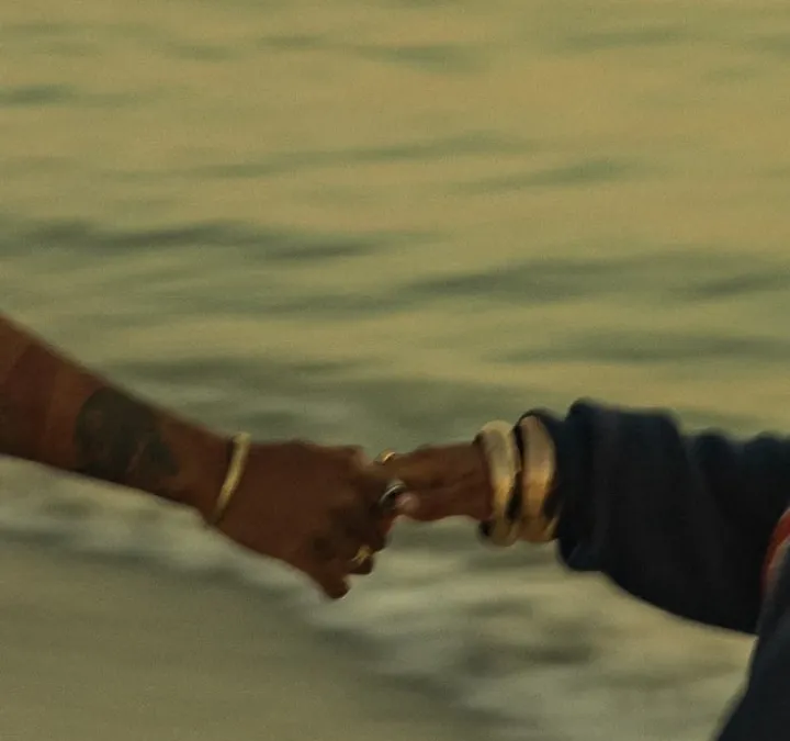 Close-up of two hands with gold rings and bracelets reaching toward each other over water at golden hour