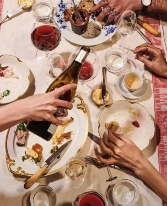 Overhead view of hands reaching across a shared dinner table with wine and gilded plates