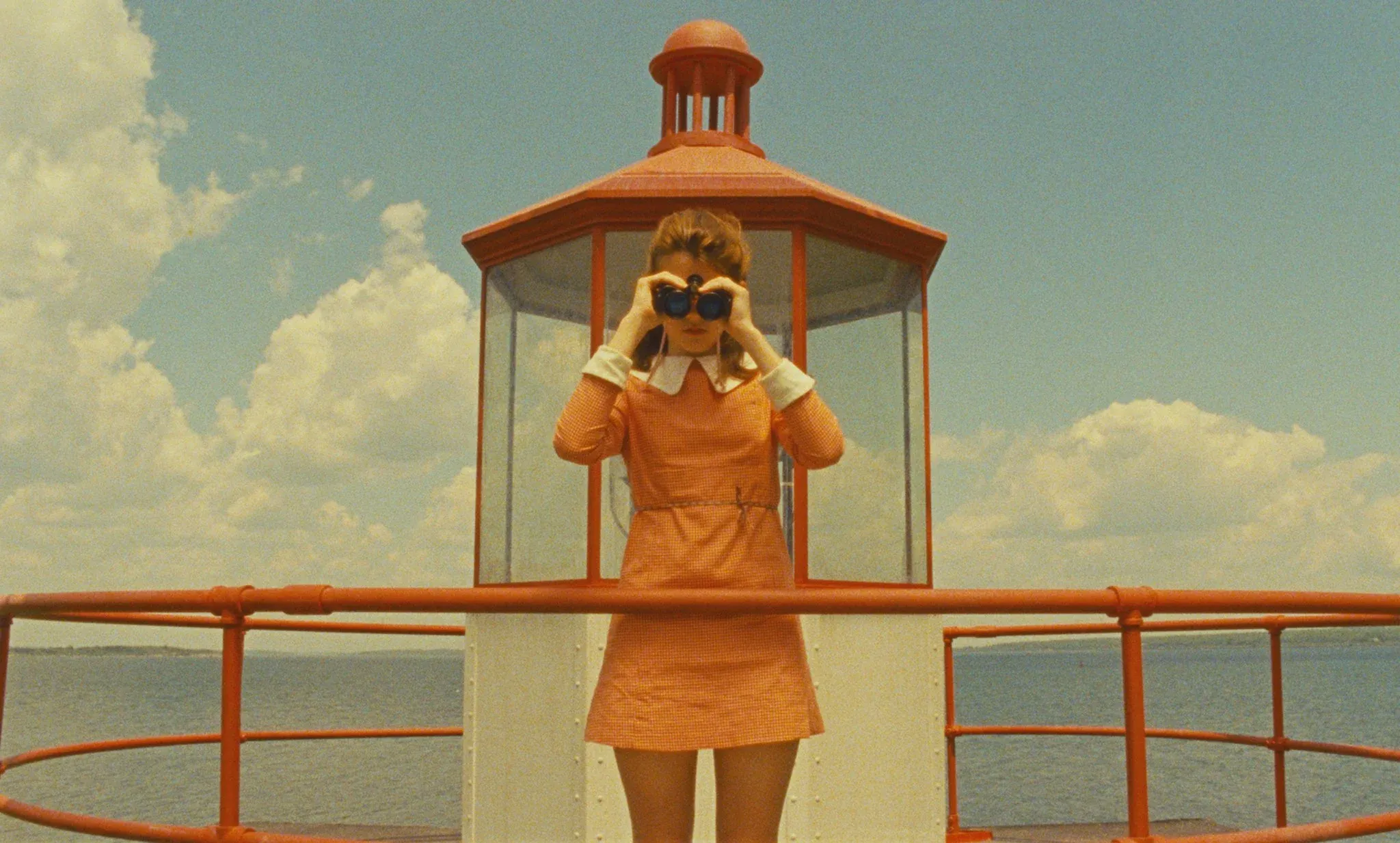 Film still of a woman in an orange dress looking through binoculars at an orange lighthouse lookout